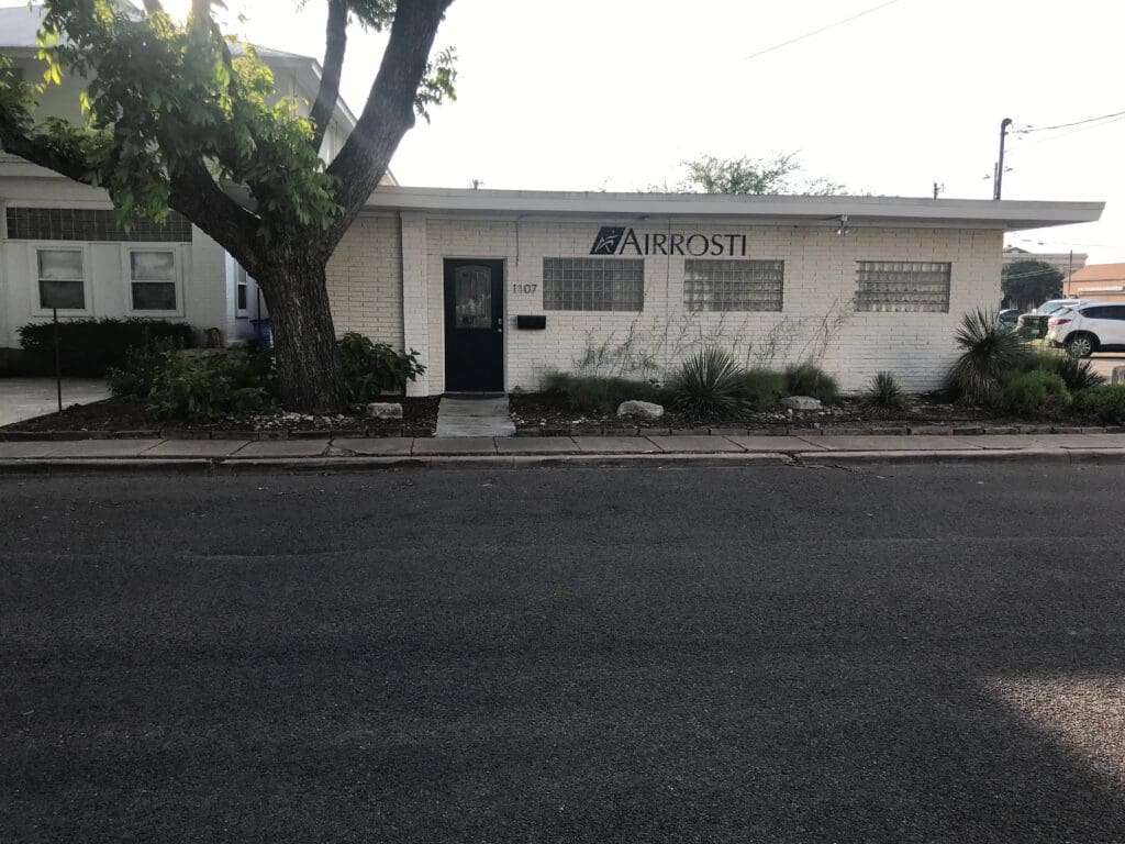 A wide view of the Airrosti Bastrop building in Bastrop, TX, just east of Austin, TX. It is a single story white brick building with the Airrosti logo displayed on the side.