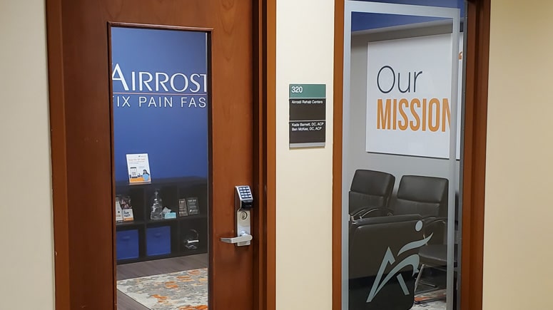 photo of the interior front entrance at Airrosti West Plano. The door is glass with the Airrosti logo on it and you can see the practice lobby through the window