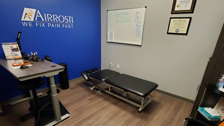 a photo of the treatment room at Airrosti West Plano. Includes the treatment table and the Airrosti logo decorates the blue painted wall in the background