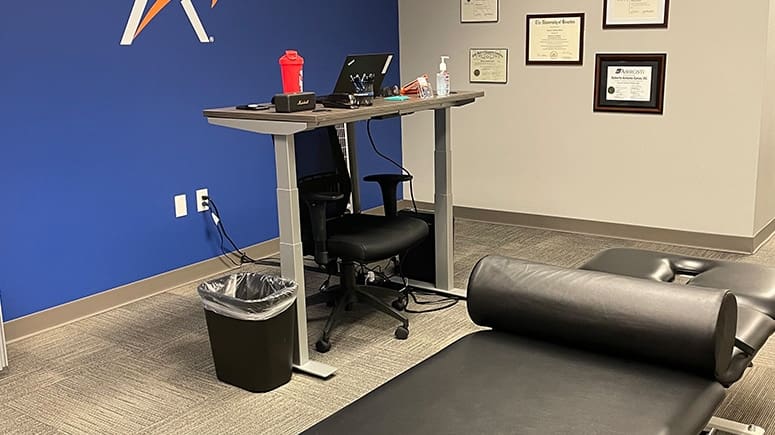 a photo of the inside of the treatment room at Airrosti Downtown Tunnels. The photo includes the treatment table, the provider's stand up desk, and the provider's accolades decorate the back wall