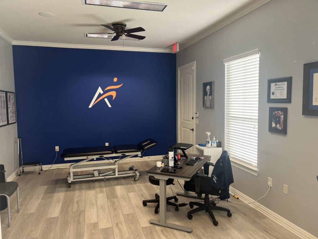 An interior view of the treatment room at Airrosti South Fort Worth. Includes the provider's desk and the treatment table. The Airrosti logo is visible on back wall.