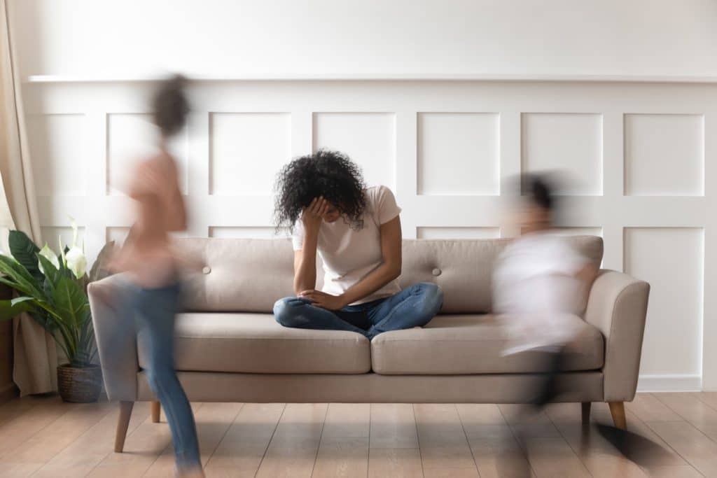 Stressed woman sitting on couch holding her head while people run in front of her