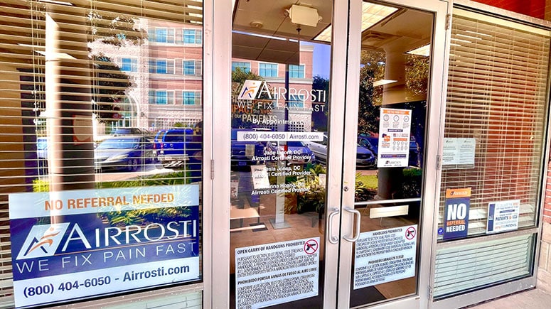 Photo of the exterior front doors at Airrosti Sugar Land. The airrosti logo and provider info are printed on the glass panes.