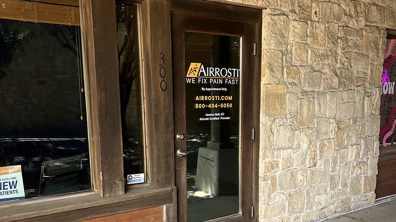 A close up view of the front door to Airrosti Cedar Hill. The Airrosti logo is displayed on the glass door, which has a wooden frame in a stone facade.